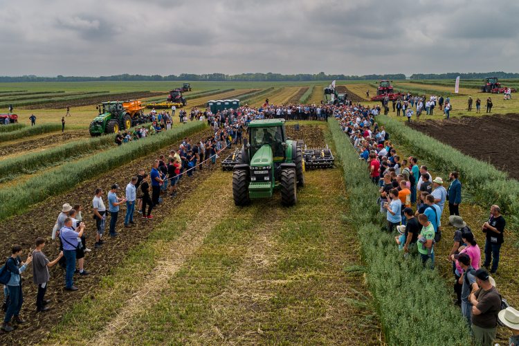 A fotón a NAK Szántóföldi Napok szántóföldi gépbemutatója látható, ahol egy traktor munkavégzés közben, nézők gyűrűjében halad a bemutatóparcellák között.